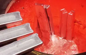 This image captures the process of melting solid aluminum. Several aluminum ingots are being added to a large crucible containing a bath of already molten, red-hot aluminum. This is a key step in both primary production and recycling.