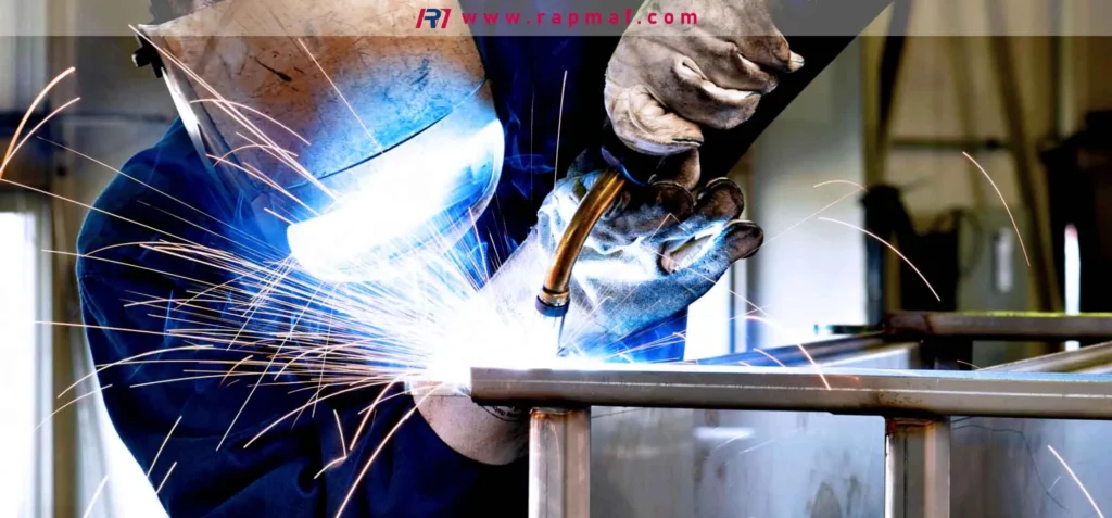 A welder wearing a protective helmet and gloves performing an arc welding process on a steel frame, creating a bright blue arc and a shower of sparks. This image represents the high-heat, base-metal-melting nature of welding, as discussed in the Rapmaf guide.