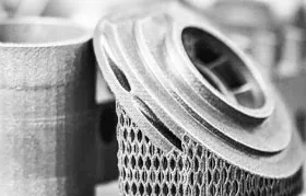 Uma fotografia em preto e branco, em close-up, de um componente industrial metálico impresso em 3D com uma estrutura de malha fina ou treliça, demonstrando uma vantagem fundamental da manufatura aditiva para geometrias complexas.
