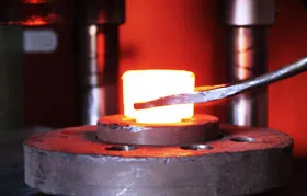 Um tarugo de aço aquecido sendo manipulado com tenazes na bigorna de uma prensa hidráulica de forjamento, imediatamente antes do golpe de prensagem que deformará plasticamente o metal no formato desejado.