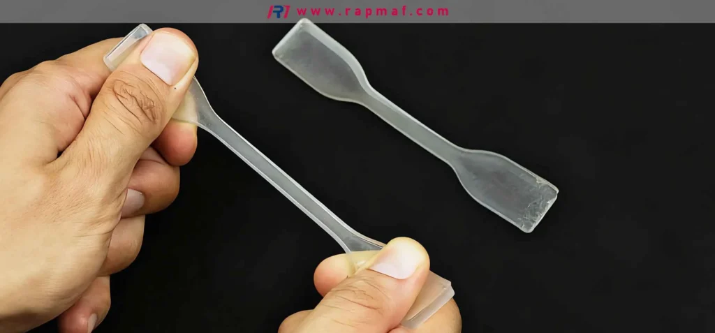 A Rapmaf image demonstrating the concept of low stiffness (low Young's Modulus). A person's hands easily bend a clear plastic 'dog-bone' tensile test specimen, showing significant elastic deformation under a small load without fracturing.