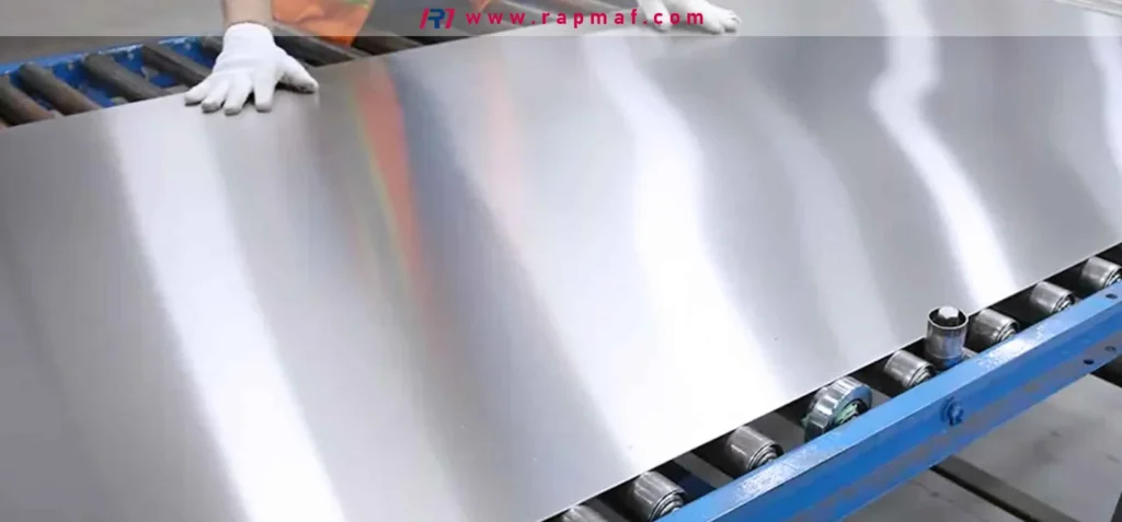 A Rapmaf image showing a worker in white gloves guiding a large, shiny sheet of metal (likely aluminum or stainless steel) along a roller conveyor in a manufacturing facility. This represents a common form of industrial metal stock used in fabrication