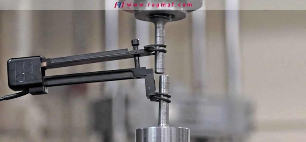 A Rapmaf image of a close-up on a tensile testing machine. A precision extensometer is clamped onto a metal 'dog bone' specimen to accurately measure its elongation under load, a standard method for determining a metal's strength, stiffness, and ductility.