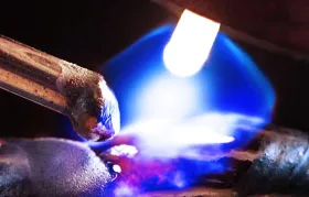 A macro photograph of an oxy-fuel torch tip with its characteristic bright blue flame, used for gas welding or brazing. In this process, gases like acetylene are used as fuel, in contrast to the inert shielding gases used in arc welding.