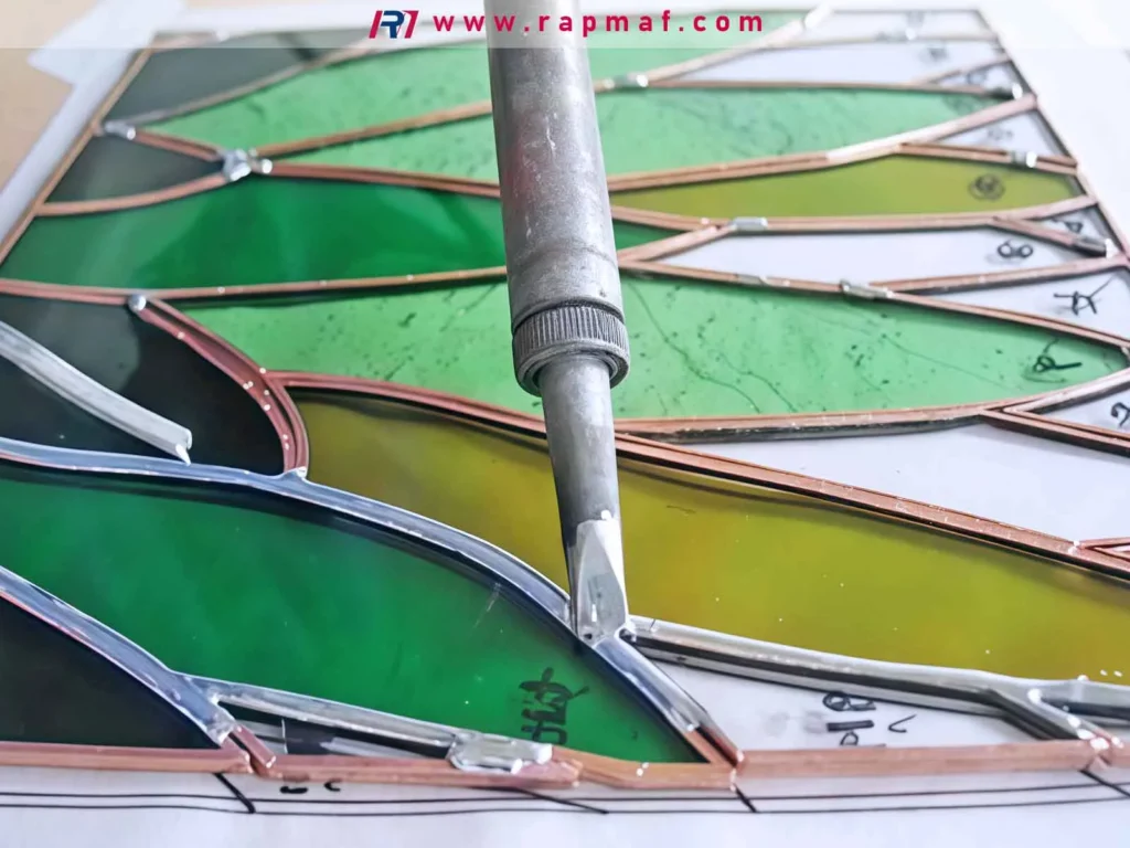 A close-up of a soldering iron being used to join pieces of stained glass with a low-melting-point solder alloy, a process related to the melting points of metals like silver and tin.