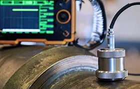 Un primer plano de un transductor de prueba ultrasónica (UT) sobre una superficie metálica curva, con la pantalla del detector de fallas en el fondo mostrando la forma de onda A-scan utilizada para el análisis de defectos.