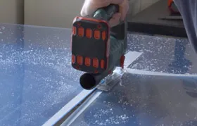 A person using a cordless jigsaw with a fine-toothed blade to make a straight cut in a clear acrylic sheet, a common method for cutting plastic without cracking or melting.