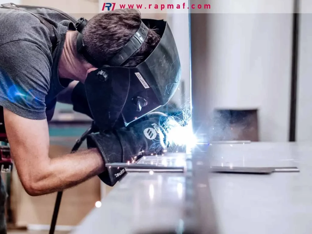 A skilled welder wearing a protective helmet and gloves performing a MIG or TIG weld on a metal workbench, an essential joining operation in metalworking that fuses parts together with an electric arc.
