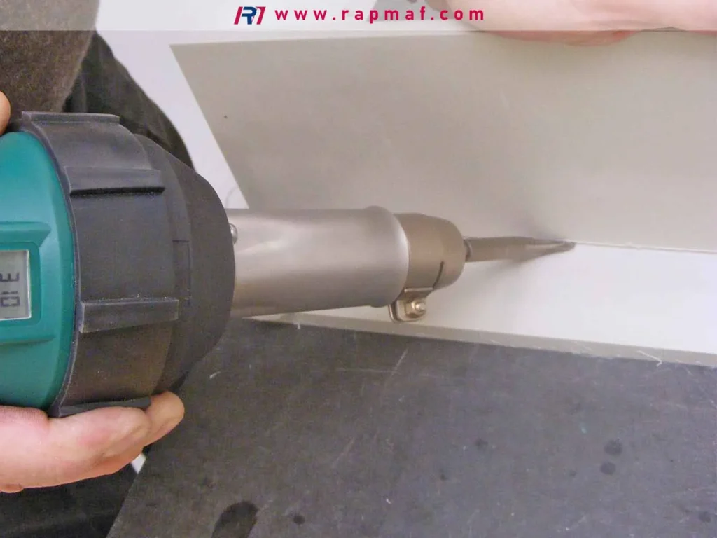 A close-up of a hot air plastic welding gun being used to fuse a seam between two white thermoplastic sheets, a common fabrication technique discussed in Rapmaf's guide.