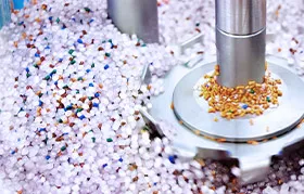 A close-up photograph of multi-colored plastic pellets (granules) being mixed in an industrial machine, representing the raw polymer resin used in manufacturing processes like injection molding and extrusion