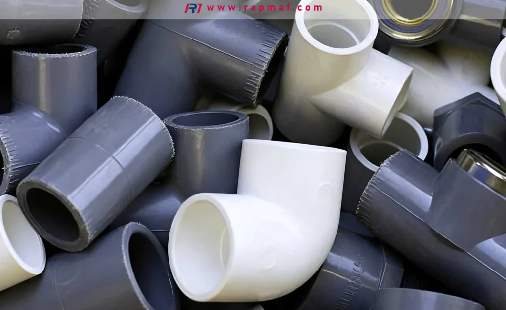 A close-up photograph by Rapmaf showing a large pile of rigid PVC pipe fittings, including gray and white elbows and connectors, a common application for polyvinyl chloride in plumbing and construction.